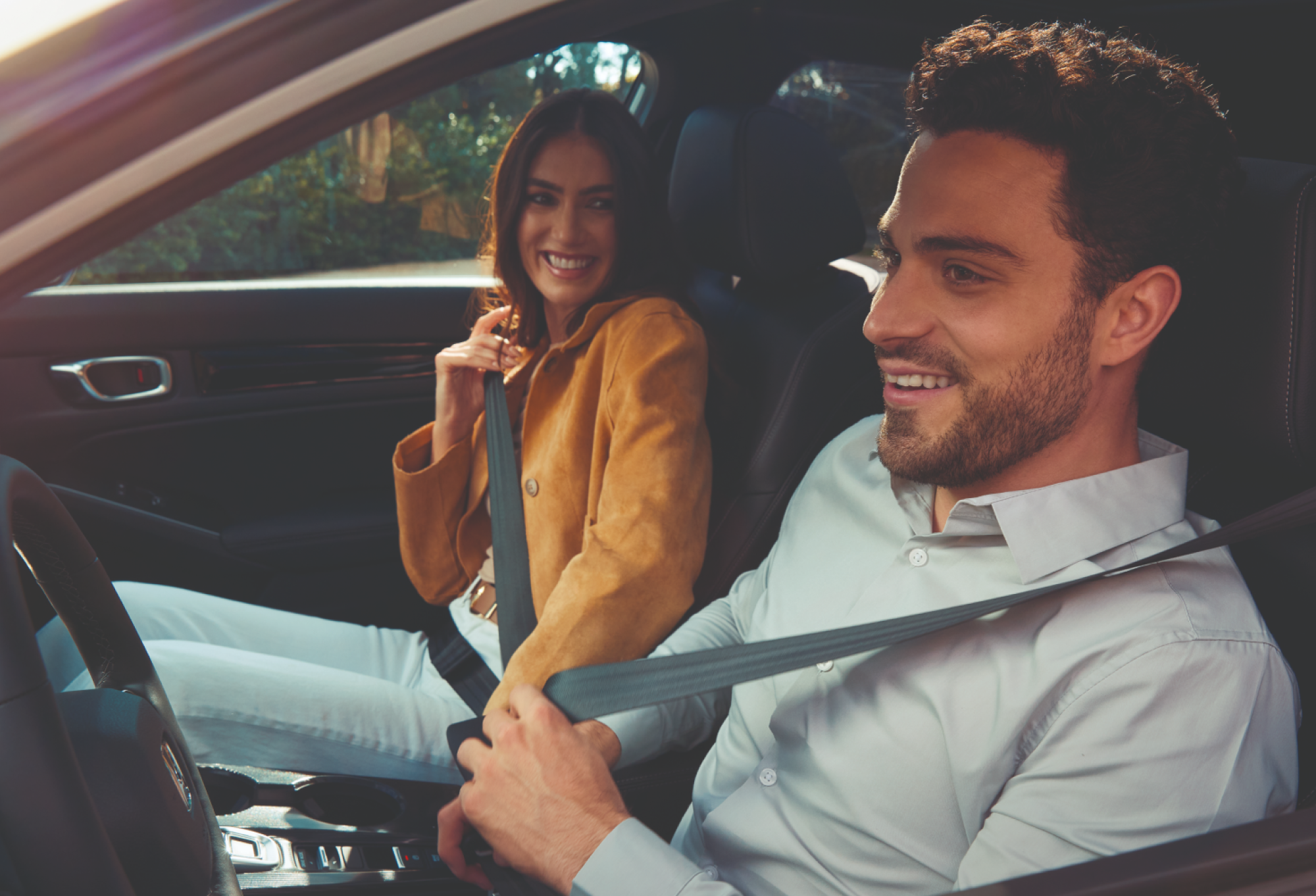 Two people sitting in the front seats of a car, smiling. The driver wears a white shirt and is fastening his seatbelt. The passenger wears a yellow jacket and is also securing her seatbelt. The interior of the car appears to be a Honda model, based on the visible dashboard design.